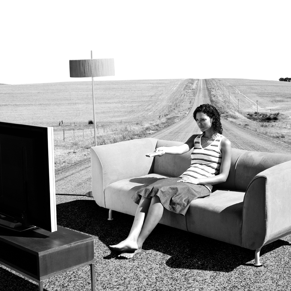 Eine Frau sitzt auf einer abgelegenen Straße auf einer Couch vor einem Fernseher mit einer Fernbedienung in der Hand. Links daneben steht eine Lampe.