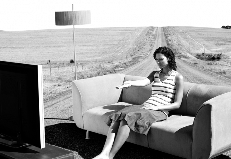 Eine Frau sitzt auf einer abgelegenen Straße auf einer Couch vor einem Fernseher mit einer Fernbedienung in der Hand. Links daneben steht eine Lampe.